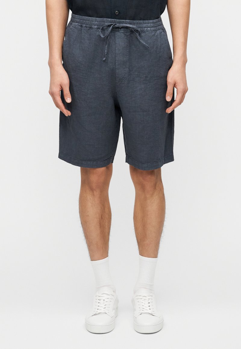 Homme portant un short gris foncé à cordon, des chaussettes blanches et des baskets blanches, debout devant un fond clair uni.