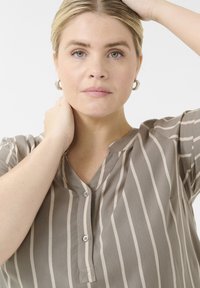 Blusa de rayas marrón y beige con escote en V, mangas cortas y cierre de botones. Textura suave con un brillo sutil y un detalle de bolsillo.
