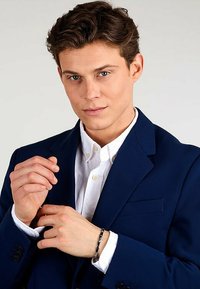 Navy blazer with notch lapels over a white button-up shirt. Silver bracelet on the wrist, model displays a neutral expression and styled hair.