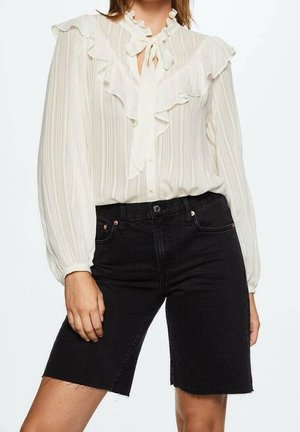 Kobieta ubrana w białą bluzkę z falbanami i długimi rękawami oraz czarne, krótkie szorty z denimu z wysokim stanem i surowymi krawędziami.