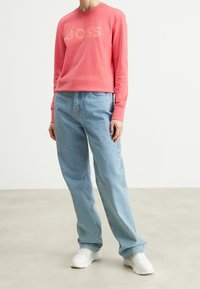 Sudadera rosa con el logotipo "BOSS", de corte relajado, cuello redondo, combinada con jeans de mezclilla de corte relajado en azul claro y zapatillas blancas.