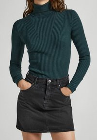 Frau in einem dunkelgrünen gerippten Rollkragenpullover und einem schwarzen Jeans-Minirock, mit den Händen in den Taschen vor einem schlichten Hintergrund.