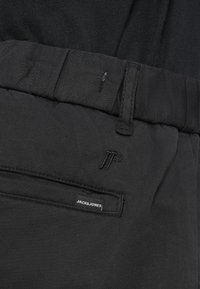 Pantalones negros con una cinturilla visible, bolsillo lateral y etiqueta de marca. Presentan detalles de costura y un logo bordado en el frente.