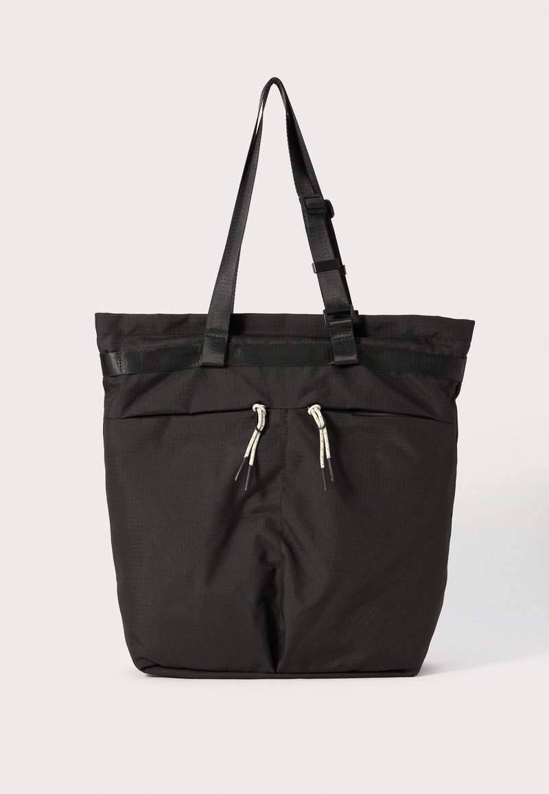 Svart tote-bag laget av slitesterk stoff, med snøring, to stropper og en teksturert overflate. Enkel design med minimale detaljer.