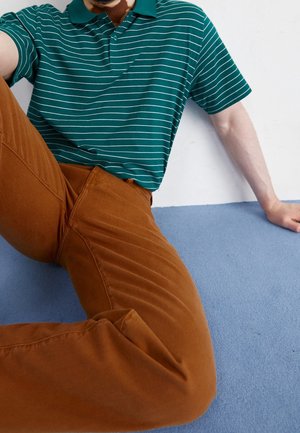 Person, der sidder på en blå tæppe iført brune bukser og en grønstribet kortærmet poloshirt, lænende sig tilbage på den ene arm.