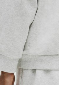 Lys grå sweatshirt med ribbede manchetter og kant, prydet med et diskret logo på siden. Blød tekstur og afslappet design.