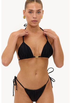 Femme portant un bikini noir à nouer avec des détails dorés en forme de soleil, tenant les liens du haut de bikini près de ses épaules.