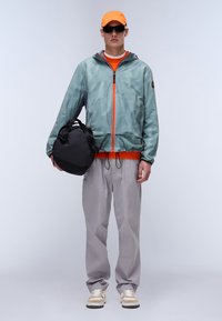 Lys grønternet jakke med orange accenter, grå bukser og beige sneakers. Holder en sort rund taske. Klar orange kasket og solbriller.