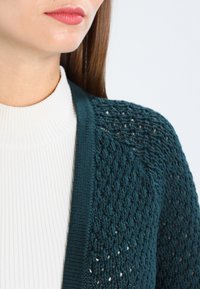 Türkiser Strickcardigan mit offener Struktur, geripptem Saum und weicher Textur, über einem weißen, gerippten Rollkragenpullover getragen.