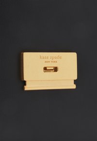 Gold-plated metal clasp with engraved "kate spade NEW YORK" text, rectangular shape, mounted on a textured black background.
