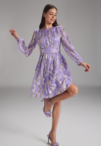 Robe lavande à manches longues, ornée de lignes ondulées dorées. Taille cintrée, jupe évasée. Portée avec des talons assortis lavandes.