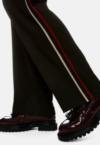 Wide-leg brown trousers with red and cream side stripes, paired with glossy burgundy shoes featuring a thick sole and tassel detail.