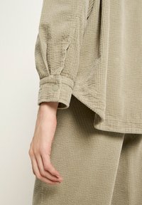 Beige corduroy long-sleeve shirt with buttoned cuff and matching pants worn on left arm and hand visible against plain background.