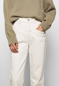 Beżowy sweter dzianinowy z prążkowanymi mankietami w połączeniu z białymi spodniami dżinsowymi w klasycznym stylu pięciu kieszeni oraz z metalowym zapięciem.