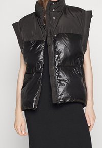 Czarna bezrękawnikowa kurtka puchowa z wysokim kołnierzem i przednimi kieszeniami, noszona na czarnej sukience z prążkowanego materiału, na modelce z luzem w ramionach.