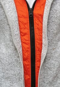 Gros plan sur des vêtements superposés avec un pull à fermeture éclair gris sur une veste matelassée orange, les deux fermetures éclair partiellement ouvertes.