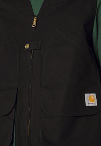 Schwarze Arbeitsweste mit einem Frontreißverschluss, zwei großen Taschen und goldenen Beschlägen. Hat ein kleines Carhartt-Logo auf der unteren rechten Tasche.