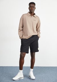 Polo de manga larga en color beige con un corte relajado, combinado con pantalones cortos negros y zapatillas blancas. Detalle notable: pequeño logo en la camisa.