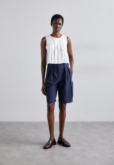 Weißes, plissiertes, ärmelloses Oberteil kombiniert mit marineblauen, gestreiften Shorts mit seitlichen Taschen, ergänzt durch burgunderfarbene Slipper mit silbernen Akzenten.