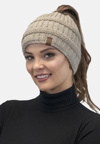 Bonnet en tricot beige avec un design côtelé et une petite étiquette logo, adapté pour accueillir une queue de cheval à l'arrière.