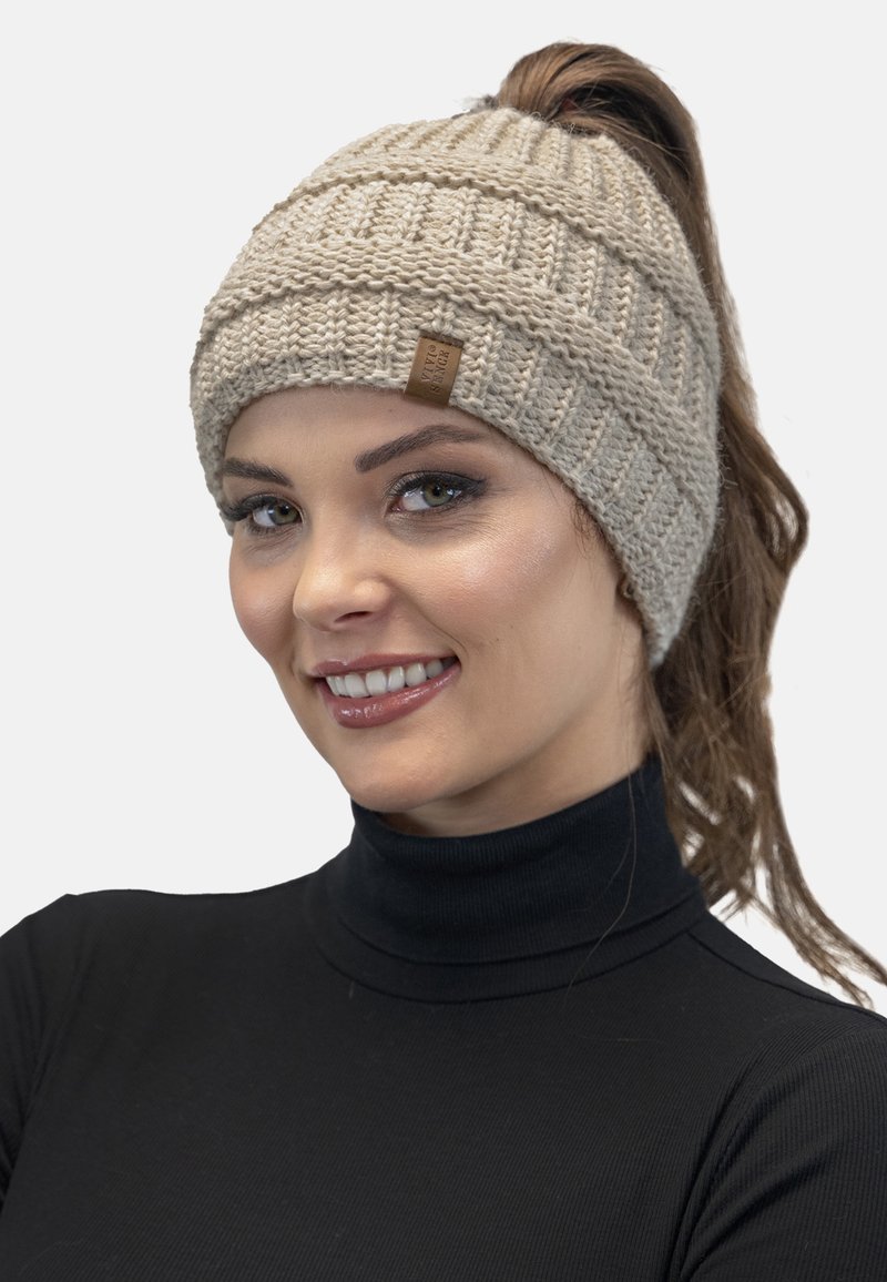 Bonnet en tricot beige avec un design côtelé et une petite étiquette logo, adapté pour accueillir une queue de cheval à l'arrière.