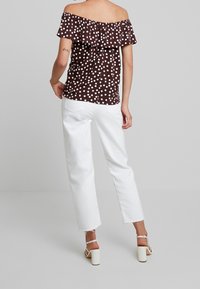 Ruskea olkapäiltä avattu paita, jossa on valkoisia pilkkuja, röyhelökaulus ja rento muotoilu, yhdistettynä valkoisiin suoralahkeisiin housuihin ja paksupohjaisiin sandaaleihin.