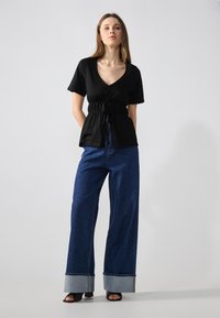 Black short-sleeve top with a drawstring waist, paired with wide-leg blue denim jeans featuring contrasting denim cuffs and open-toe black heels.