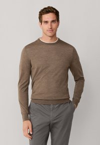 Bruin gebreid sweater met een ronde halslijn en lange mouwen. Heeft een subtiele textuur en een klein logo op de borst. In combinatie met grijze broeken.