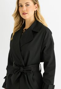 Black trench coat with a belted waist, large lapels, and button details. Fabric appears smooth. Visible animal print underneath.