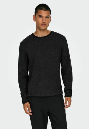 Jeune homme portant un pull texturé gris foncé à manches longues et un pantalon noir, debout devant un fond clair uni.