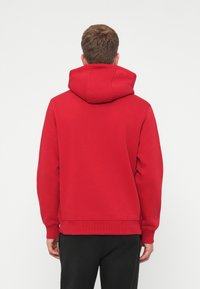Red hooded sweatshirt with a textured finish. Features a drawstring hood and ribbed cuffs. Worn with black pants, shown from the back.