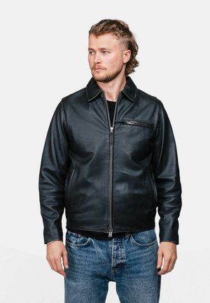 Homme avec une légère barbe et une coiffure mulet, portant une veste en cuir noire et un jean bleu, regardant sur le côté sur un fond uni.