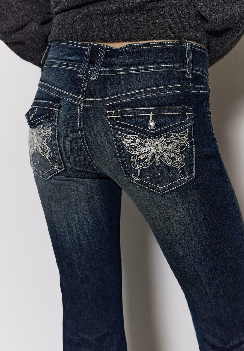 Dunkelblaue Jeans aus Denim mit verzierten Gesäßtaschen, die Schmetterlingsmuster und silberne Akzente aufweisen. Gerade geschnittener Beinverlauf mit mittelhoher Taille.
