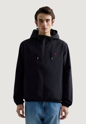 Ung mand iført en sort hættetrøje med en rød logo på brystet og blå jeans, står foran en lys grå baggrund.