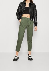 Pantalons en coton verts avec une coupe slim, ourlet à revers et poches avant, associés à une veste bomber en cuir noire et des mocassins blancs et noirs.