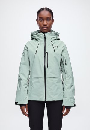 Une femme porte une veste à capuche vert pâle, avec des fermetures éclair noires et des cordons de serrage, sur un fond blanc uni.