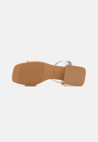 Transparent sandal med klack, fyrkantig tå, gummisula med texturerat mönster och justerbar spänne.