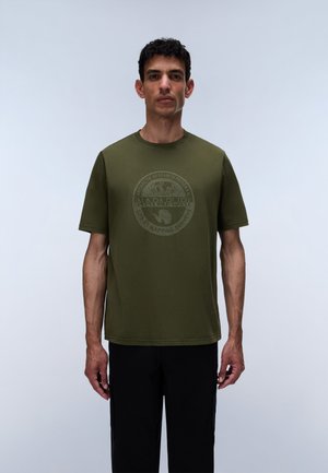 Man standing against light background wearing an olive green short-sleeve T-shirt with a circular Antarctic research logo.