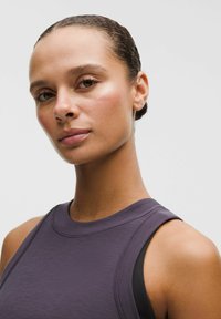 Top sportiv fără mâneci, de culoare mov, cu decolteu rotund. Material neted, ușor, cu detalii de cusătură; purtat peste un top sport negru.
