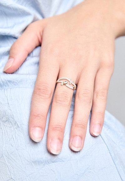 Ruka nosi zlatý prsteň s malými diamantmi a dizajnom včely, ktorý spočíva na svetlomodrej textúrovanej látke.