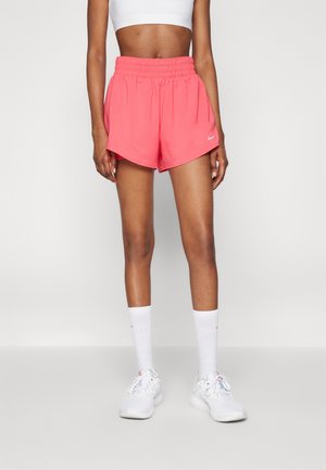 Vrouw die koraalroze Nike sportshorts draagt, een witte sportbh, witte hoge sokken en witte sneakers, staand tegen een egale achtergrond.