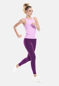 Junge Frau joggt im Mittelschritt, trägt ein pinkfarbenes ärmelloses Oberteil, lila Leggings und weiße Turnschuhe vor einem einfarbigen Hintergrund.