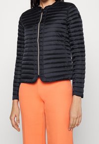 Black puffer jacket featuring horizontal quilted sections, a round neckline, and a front zipper. Worn with bright orange wide-leg pants.