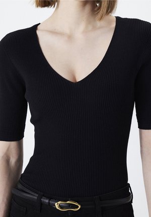 Femme portant un haut noir côtelé à col en V et manches courtes, rentré dans un pantalon noir avec une ceinture noire avec une boucle dorée.
