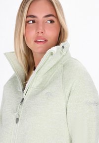 Donna con lunghi capelli biondi che indossa una giacca leggera verde chiaro con cappuccio foderato in pile, che guarda di lato su uno sfondo bianco.
