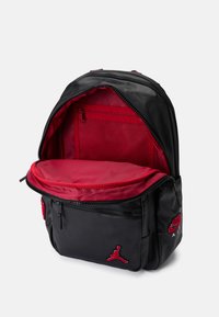 Mochila preta com interior vermelho, apresentando um compartimento principal com fecho éclair, um bolso frontal e logótipos bordados. Textura suave e material durável.