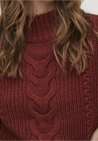 Ruskot, kihara hiukset tummanpunaisessa paksussa neulotussa villapaidassa, jossa on korkea ribbikaulus ja keskellä kantattu kuvio.