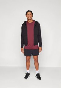 Camisola preta com capuz e fecho de correr sobre uma camisa bordô, combinada com calções azul-marinho e ténis pretos. Tecido liso com fecho de correr frontal e bolsos.