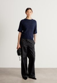 Πλεκτό πουλόβερ κοντομάνικο σε σκούρο μπλε συνδυασμένο με άνετο μαύρο παντελόνι. Το μοντέλο κρατά μια μικρή μαύρη τσάντα χειρός. Ομαλή υφή, άνετη εφαρμογή.