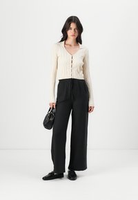 Crème gebreide cardigan met V-hals en knoopsluiting in combinatie met een wijdere zwarte broek. Accessoires bestaan uit een kleine zwarte handtas. Zwarte schoenen.